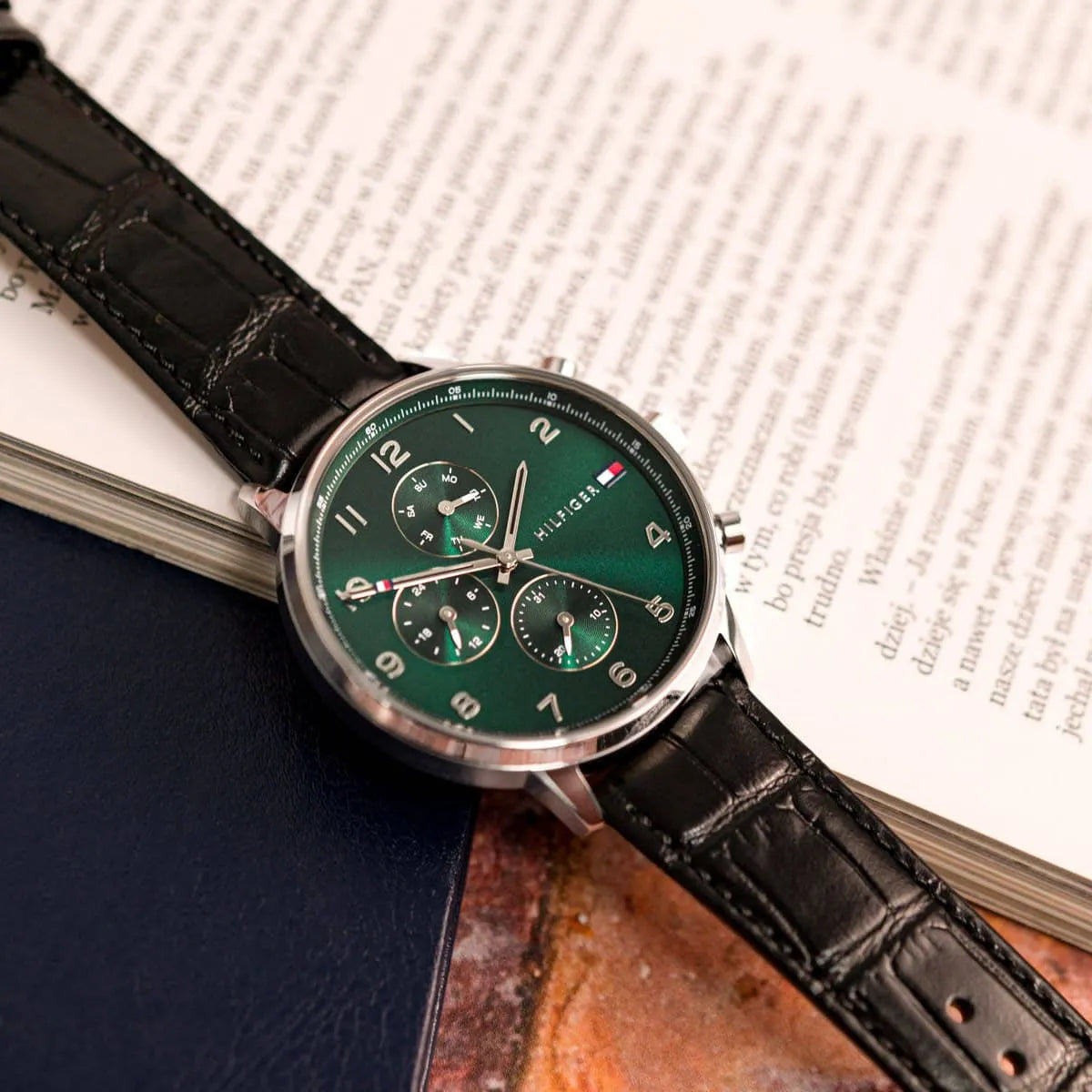 Tommy Hilfiger green dial watch on book, showing watch face and black crocodile-textured leather strap