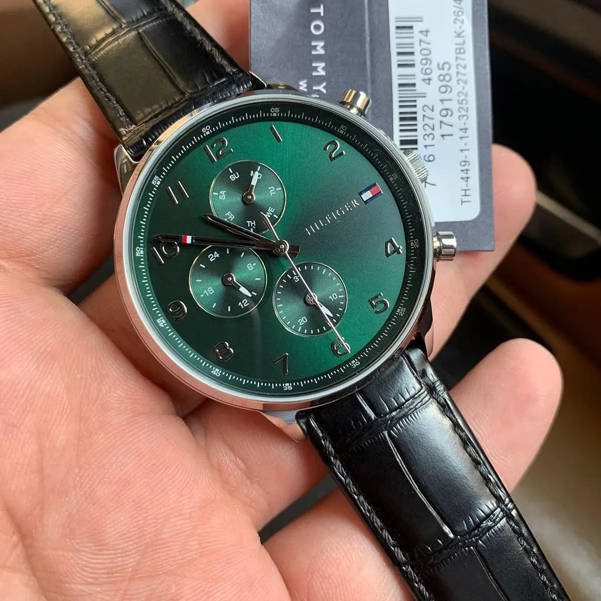 Tommy Hilfiger chronograph watch worn on wrist, displaying green dial and black leather strap with product tag