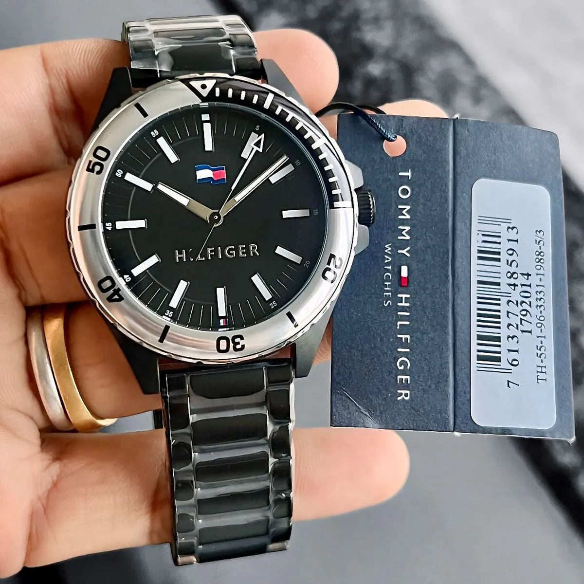 Tommy Hilfiger Logan watch on wrist with product tag, showing black dial and metal bracelet