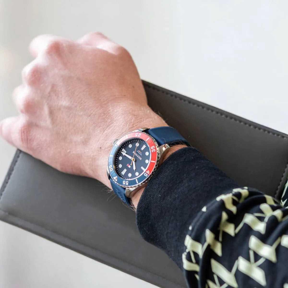 Tommy Hilfiger watch worn on wrist with blue dial, red bezel, and navy silicone strap against neutral background