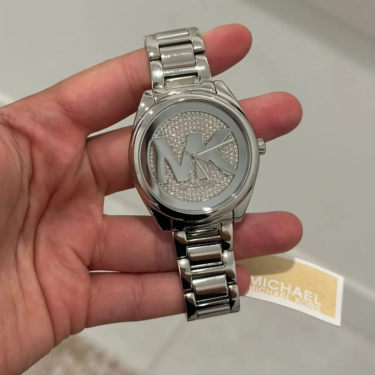 Michael Kors Janelle women's watch held in hand displaying silver dial and stainless steel bracelet with tag