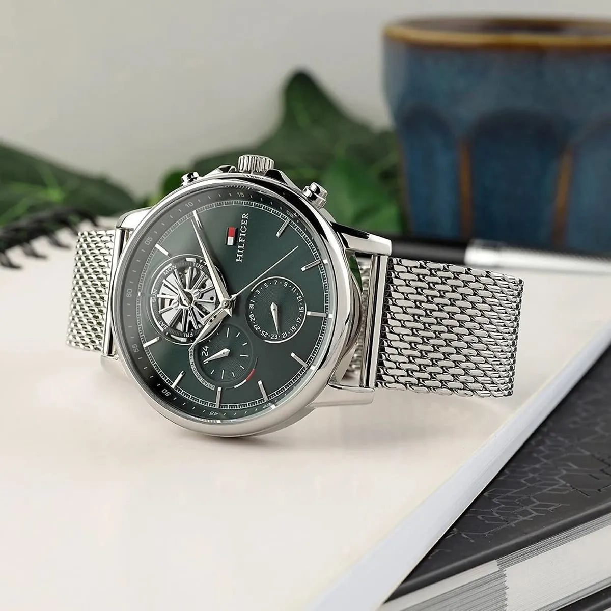 Tommy Hilfiger green dial watch with silver mesh strap on desk with decorative items