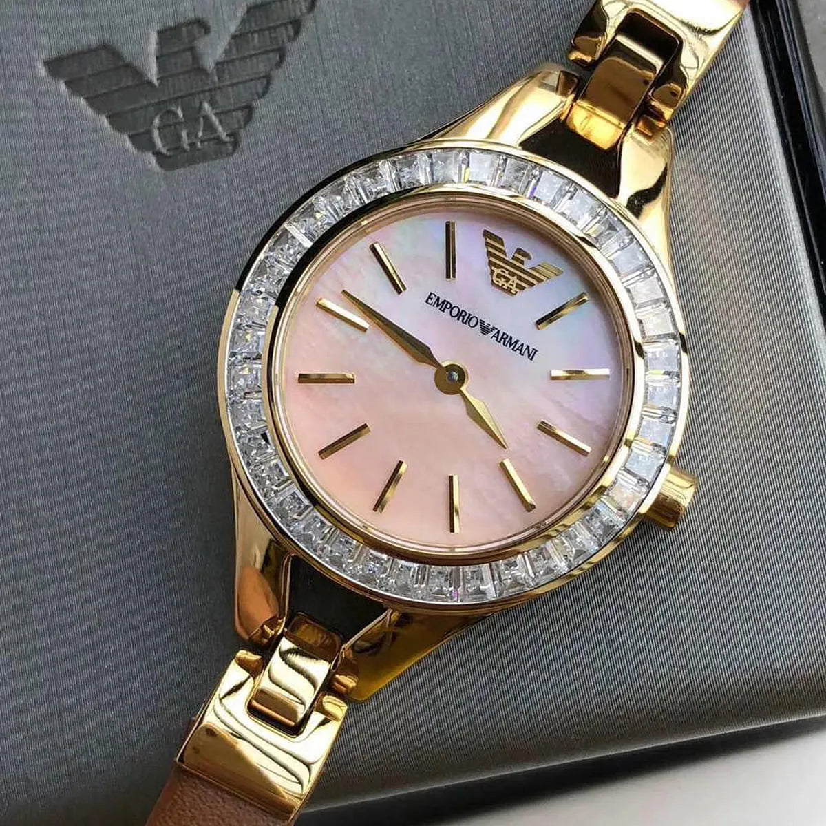 Angled view of Emporio Armani Chiara watch with gold-tone case and crystal-set bezel on Armani logo background